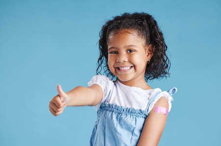 Happy vaccinated kid showing thumbs with plaster on arm after vaccine injection standing against a blue studio background. Advertising vaccination against coronavirus. Child immunisationの写真素材
