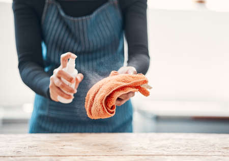 Closeup of one woman spraying antibacterial cleaner from a bottle onto a cloth to disinfect and wipe table in a cafe or store. Hands of a shop assistant sanitising surfaces to maintain hygiene and prevent the spread of covid germs.の写真素材