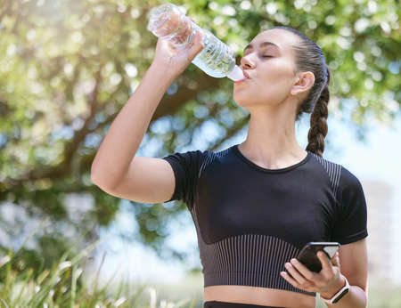 Young female athlete taking a break and drinking water from a bottle and holding smartphone while out for a run and exercising outdoors during the dayの写真素材