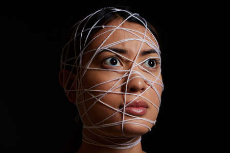 Helpless and afraid. a young woman wrapped in string against a dark background.