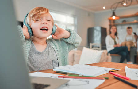 A cute caucasian little blonde girl wearing wireless headphones while being creative by drawing in a book at home with her family in the background. Girl enjoying magic of music ignoring homeworkの写真素材