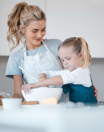Happy mother helping her child bake. Woman looking at her daughter bake. Caucasian little girl mixing a bowl of batter. Proud mother watching her daughter bake. Small girl enjoying bakingの写真素材