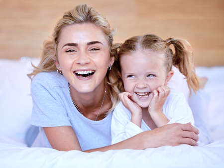 Happy caucasian mother and daughter lying on a bed at home. Cheerful woman with cute little girl enjoying a cosy and lazy relaxing day together. Loving parent bonding and sharing quality time with kidの写真素材