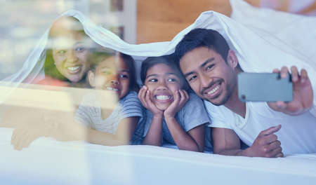 Happy parents with little children lying on bed at home under blanket and taking selfies on...