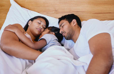 Mixed race family of three asleep in bed. Above mother, father and daughter resting on a white bed in the bedroom at home. Tired parents lying in bed sleeping with their daughter in the middleの写真素材