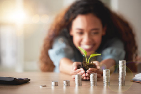 African American woman holding a growing plant in her hands while counting her savings. Stacks of...