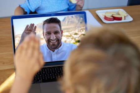 Closeup of a son having a video call with his caucasian father on a laptop at home and greeting with a waving gestureの写真素材