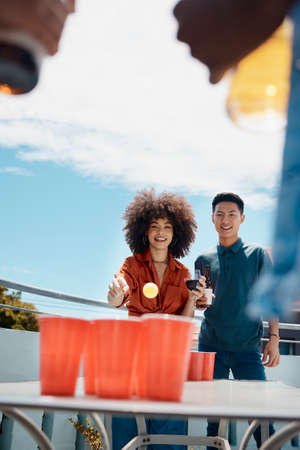 Happy young friends playing beer pong together outside. Diverse friends throwing a ball into cups of...