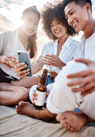 A group three multiracial friends relaxing and using a phone at the beach while having alcoholic drinksの写真素材