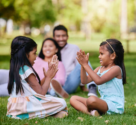 A happy asian couple lying together on grass outside, loving parents enjoy quality time with their little daughters playing a game. Couple bonding during family time at park with adopted foster sisterの写真素材