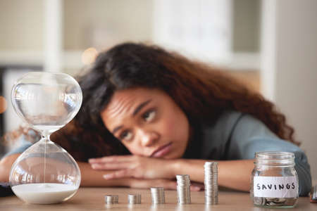Young african american woman staring at an hour glass while looking bored at home. Mixed race person counting down while financial planning in her living room. Waiting for her investments and savingsの写真素材