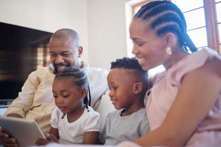 A young african american family sitting on the sofa together and smiling while using a tablet at...