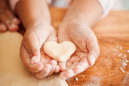 Closeup of an unknown chid holding a heart shaped cookie while baking in the kitchen with her mother. An unrecognizable mother teaching her daughter domestic skills at homeの写真素材