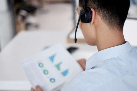 Businessman working in a call center reading a document. Call center operator wearing a headset. Customer service rep reading paperwork. Businessman looking at finance reports. Marketing rep on a callの写真素材