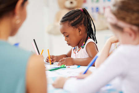 African american girl sitting at a table and colouring at pre-school or kindergarten with her fellow students. Young female child using colourful pencils to draw pictures in a classroom at her schoolの写真素材
