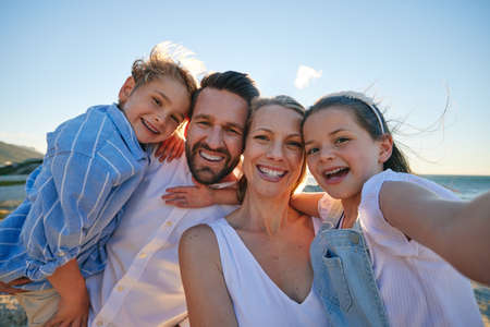 Caucasian taking a selfie on the beach. Happy family on holiday taking a photo by the ocean. Portrait of a cheerful family on vacation. Young mother and father bonding with their children.の写真素材