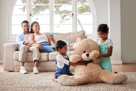 Young couple relaxing on a sofa while their two girls play on the living room floor with their soft teddy at homeの写真素材