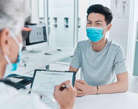 Filling out the mandatory form. a young man wearing a face mask and sitting with his doctor during...