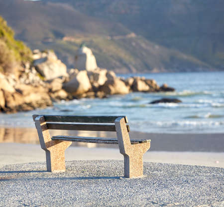 Beautiful relaxing view of the ocean with a bench at the beach on a summer day. The landscape of the sea shore with big rocks and calm waves on a peaceful spring afternoon on the coastlineの写真素材