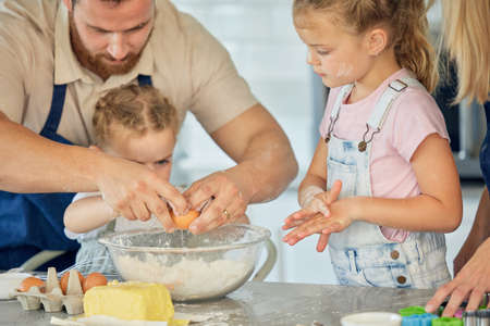 Caucasian father helping little daughter crack an egg into a bowl while baking in the kitchen at...