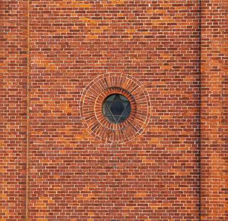 Wall of a jewish synagogue with round window on an old red brick house outside. Small metal frame on historic temple building with Star of David design and architectural background with copy spaceの写真素材