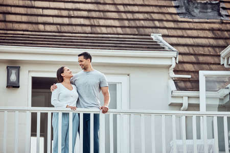 Happy young mixed race couple standing on the balcony at their new home. Hispanic couple looking into each others eyes. Loving husband and wife standing outside getting fresh airの写真素材