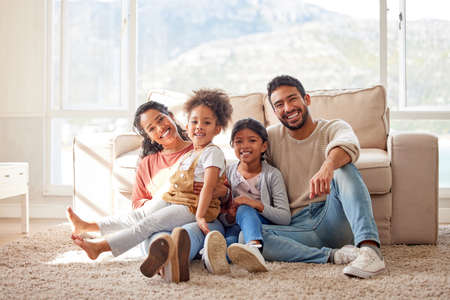 Portrait of two happy parents bonding with their daughters in the lounge, sitting on the floor. Smiling young family relaxing at home, enjoying time together. Mother and father holding their daughterの写真素材