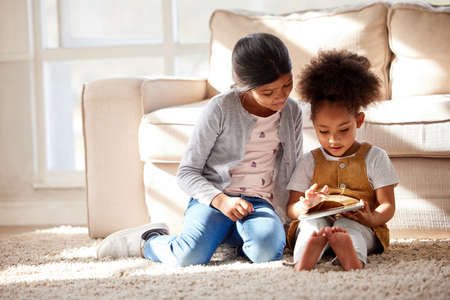 Two little mixed race sisters sitting on the floor and playing on digital tablet together at home....