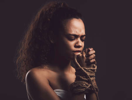 a young woman looking scared with a rope tied around her arms against a black background.