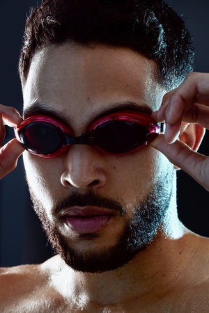 Water you waiting for Lets dive in. Studio shot of a fit young professional swimmer putting on his goggles against a dark background.の写真素材