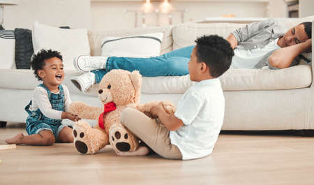 Dad needs a break. two siblings fighting over a teddy on the floor while their dad sleeps on the couch at home.の写真素材