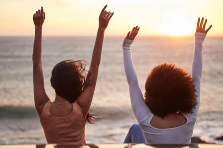 Love is a gift of the gun. Rearview shot of two young unrecognizable females celebrating while watching the sunset at the beach.の写真素材