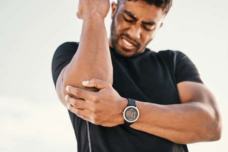How do I exercise now. a handsome young male athlete holding his elbow in pain while exercising on the beach.の写真素材