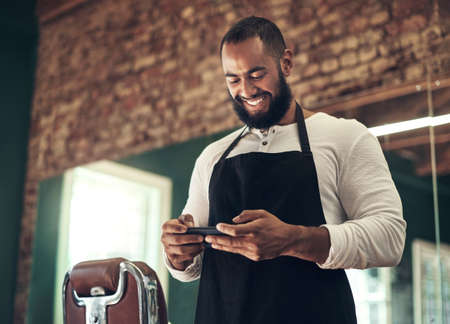 Keeping up with the new hair trends. a handsome young barber standing alone in his salon and using his cellphone.の写真素材