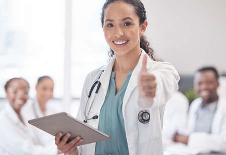 Youve got my backing. Cropped portrait of an attractive young female doctor giving thumbs up and using a tablet while standing in the boardroom with her colleagues in the background.の写真素材