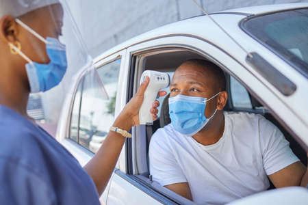 Drive thru for covid screening or testing service for people driving or traveling. Black man wearing a mask in a car getting his temperature test using an infrared thermometer as coronavirus protocolの写真素材