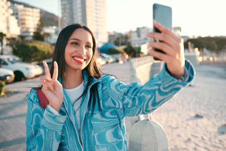 Fun, happy and trendy student taking a selfie on phone for social media while exploring, visiting or enjoying city. Stylish, edgy and funky woman showing peace sign, symbol and gesture for social appの写真素材