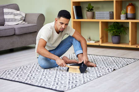 Scoop it all up. a young man using a dustpan and sweeping his carpet at home.