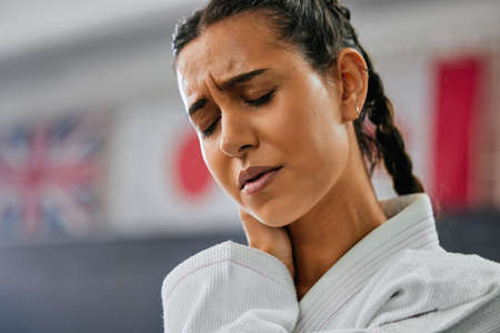 Young active female with pain from training in a karate and fitness studio. Portrait of a fit woman with a head and neck injury from a fight class and workout. Closeup of a tired face after exerciseの写真素材
