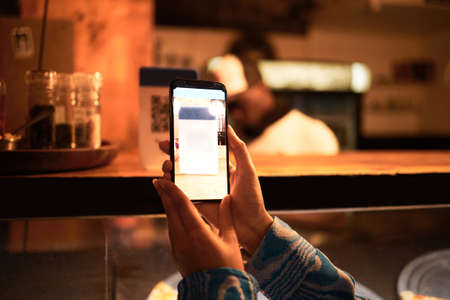 Person with phone camera scanning QR bar code for online menu at a modern restaurant, shop or cafe at night. Hand of a woman taking photo scan for website information using modern technologyの写真素材