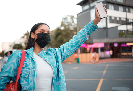 Trendy, stylish and funky student with phone travel app wearing mask and hailing, or waving car, taxi and local transport or lift service. Cool, fashionable and careful woman in cityの写真素材