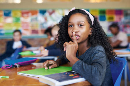 Shh, youre not supposed to talk. a young girl sitting in her school classroom with her finger on her lips in a hushing gesture.の写真素材