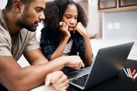 Class by day, home by night. a young girl using a laptop while doing a school assignment with her father at home.の写真素材