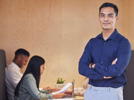 My team ensures success. Cropped portrait of a handsome young businessman standing with his arms crossed in the boardroom with his colleagues in the background.の写真素材