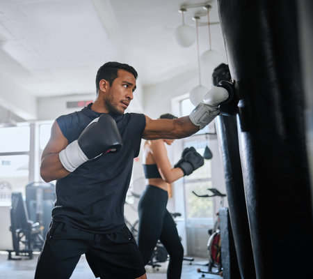 Active, fit and serious man boxing, sweating and doing a cardio workout at the gym. One sporty, sweaty and determined male athlete looking tired while punching a bag at a sports center for fitnessの写真素材