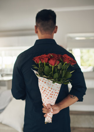 Can you guess whats behind my back. a young man holding flowers behind his back at home.の写真素材