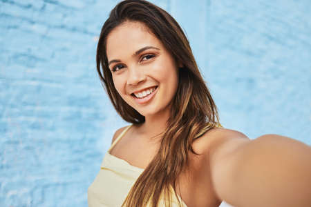 Summer selfies. Cropped portrait of an attractive young woman taking selfies outside against a blue background.の写真素材