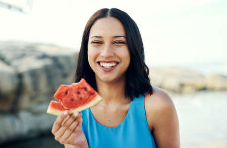 Hmm nothing beats fresh watermelon. a young woman eating a slice of watermelon on the beach.の写真素材