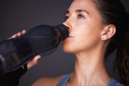 Hydration is key for peak performance. a sporty young woman drinking water against a grey...