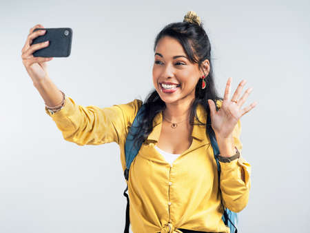 Collect moments not things. Studio shot of a woman wearing a backpack and taking selfies against a white background.の写真素材